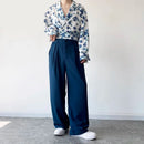 Person wearing a floral blouse and wide-leg pants against a white wall.