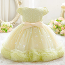 A pale yellow-green children's party dress with a ruffled tulle skirt, flutter sleeves, and a large bow at the waist, displayed against a soft indoor background with flowers and a mirror, Obroi