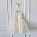 A sleeveless cream-colored children's party dress with a structured waist and a voluminous tulle tutu skirt, hanging on a wooden hanger from a decorative white wall hook, Obroi
