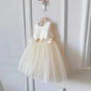 A sleeveless baby girl's party dress with a white satin bodice, bow details, and a voluminous cream tulle tutu skirt, hanging on a wooden hanger against a white paneled wall, Obroi