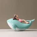 A whimsical whale-shaped chair or sculpture in mint green and white, with a small girl figurine reclining inside on a plain studio background, Obroi