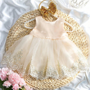 A toddler girl's formal dress in pale blush pink, featuring a satin bodice, layered tulle skirt with white floral lace trim, and a pearl-embellished waistband. The dress is styled on a woven straw mat with a gold glitter bow, pink flowers, and white satin fabric, Obroi