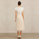 A woman viewed from the back wearing a sleeveless midi dress with a sheer lace back and a pleated tulle skirt in a light cream or off-white shade, standing against a neutral textured beige wall, Obroi
