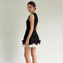 A woman modeling a sleeveless black dress with a structured back, a white ruffled underskirt peeking out at the hem, and black bow details at the bottom corners, set against a plain light gray studio background, Obroi