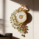 A decorative wall clock shaped like a peacock, with a gold-tone frame, white dial, and cascading peacock-feather details in iridescent blue-green and clear teardrop crystals, mounted on a beige wall with strong sunlight and shadows, Obroi