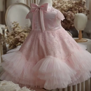 A light pink girl's party dress displayed on a mannequin, featuring a lace bodice with a large satin bow, short lace sleeves, and a layered tulle skirt, Obroi