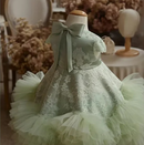 A pale mint green children's dress displayed on a dress form mannequin, featuring a lace bodice with floral embroidery, a large satin bow at the back of the neck, short puff sleeves, and a layered tulle skirt, Obroi