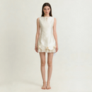 A woman stands barefoot against a plain light background wearing a sleeveless cream-colored mini dress with a tie-neck detail and decorative ruffle trim with fabric flowers at the hem, Obroi