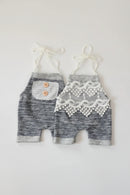 A pair of baby shorts in heathered gray and white, featuring one with a white lace pocket with two wooden buttons and the other with a white lace panel, both with white drawstrings, laid flat on a white background, Obroi