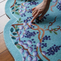 Hand touching a floral patterned rug on a wooden floor, Obroi