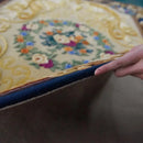 Hand holding a blue mat over a decorative rug with floral patterns, Obroi