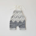 Two baby romper shorts in heathered gray fabric, laid flat on a white background. One has a white textured pocket with two wooden buttons and white drawstrings, and the other has a white lace panel and white drawstrings, Obroi