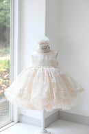 A sleeveless cream-colored toddler girl's party dress with a lace bodice, tulle skirt, and matching flower crown, displayed on a mannequin in front of a window, Obroi