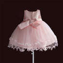 A light pink sleeveless party dress with a large bow at the waist and a layered tulle skirt with a ruffled lace hem, displayed on a stand against a dark brown background, Obroi