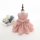 A light pink lace toddler dress displayed on a mannequin dress form, featuring a sleeveless bodice, flared skirt, and a large matching bow at the waist. A small floral arrangement sits to the left on a white surface against a white background, Obroi
