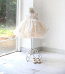 A cream-colored lace and tulle toddler party dress displayed on a white dress form stand, with a matching headband and small shoes on a marble floor next to a window, Obroi