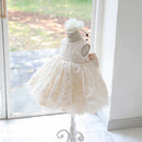 A sleeveless baby girl's dress in cream lace and tulle, displayed on a white mannequin dress form with a matching headband, set in a bright room near a window, Obroi