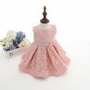 A pink lace toddler girl's dress displayed on a small dress form mannequin, featuring a sleeveless bodice, full skirt, and a rhinestone-embellished waistband with a pink bow. A small floral arrangement in a white box sits to the left on a white background, Obroi
