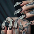 Close-up of a hand with long, pointed nails and metallic accents against a dark background, Obroi