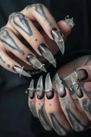 Close-up of tattooed hands wearing long, sculpted coffin-shaped press-on nails with a smoky translucent gray and black gradient and small metallic ring and spike embellishments, Obroi