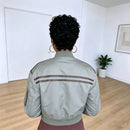 A person viewed from behind wearing a light sage green bomber jacket with brown stripes on the shoulders, standing in a bright room with light wood floors and white walls, Obroi