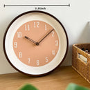 Round wall clock with a peach-pink face, dark brown rim, and white numerals, shown on a wooden surface against a light wall with a size marker reading 30cm/11.8inch above, Obroi
