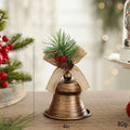 A decorative Christmas bell ornament with a distressed bronze finish, green pine needles, red berries, and a burlap ribbon loop, shown on a tabletop with size markings of 10cm, 8cm, and 80g, Obroi