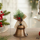 A decorative Christmas bell ornament with a distressed bronze finish, green pine needles, red berries, and a burlap ribbon loop, shown on a tabletop with size markings of 10cm, 8cm, and 80g, Obroi