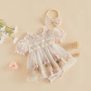 A baby girl's short-sleeve romper dress in cream/off-white tulle and mesh, decorated with pink and green floral embroidery, lace trim, and a pearl-like chest band. It is styled in a flat-lay with a matching cream bow headband, white flowers, and small decorative balls on a solid beige background, Obroi