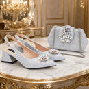 A pair of sparkling silver high-heeled pumps with matching embellished handbag, displayed on a marble table in an ornate gold-accented room, Obroi
