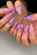 A close-up of hands displaying a set of long, almond-shaped press-on nails with a pink and purple celestial design, including swirls, stars, and rhinestone accents, Obroi