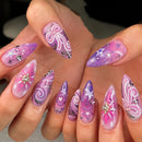 A close-up of hands displaying a set of long, almond-shaped press-on nails with a pink and purple celestial design, including swirls, stars, and rhinestone accents, Obroi