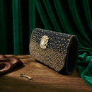 A black evening clutch bag heavily embellished with gold studs and a large ornate gold crest clasp, resting on a wooden table with a green velvet curtain backdrop, Obroi