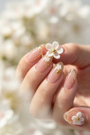 A close-up of a hand showcasing elegant almond-shaped nails with a glossy nude-pink base, decorated with delicate gold leaf swirls, tiny white flower charms, and small green accents. A larger white flower with a gold center is held near the middle finger against a soft, blurred background of similar light tones, Obroi