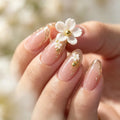 A close-up of a hand showcasing elegant almond-shaped nails with a glossy nude-pink base, decorated with delicate gold leaf swirls, tiny white flower charms, and small green accents. A larger white flower with a gold center is held near the middle finger against a soft, blurred background of similar light tones, Obroi