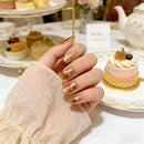 Hand with gold glittery nails holding a small dessert, with a tea set and pastries in the background, Obroi