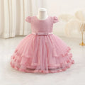 A pink toddler girl's party dress displayed on a white mannequin dress form. The dress features a textured honeycomb lace bodice, short puff sleeves, a matching pink satin waistband, and a tiered tulle skirt with lace trim, Obroi
