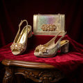 Gold high-heeled shoes with embellishments and a matching handbag on a red fabric background, Obroi