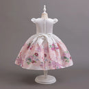 A white girl's party dress displayed on a white mannequin, featuring a floral pink and purple patterned skirt with a large white bow at the waist and puffed sleeves, Obroi