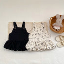 Two children's dresses, one black and one floral, on a textured surface with a basket and shoes in the background, Obroi