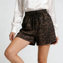 A person wearing leopard-print shorts with a black drawstring waist, paired with a white satin blouse, shown against a light gray studio background, Obroi