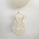 A cream-colored baby romper with lace trim and wide shoulder straps, hanging on a decorative pearl-bead hanger beneath a matching paper lantern against a plain white wall, Obroi