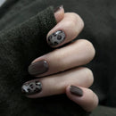 Obroi Hand with dark brown nail polish and black leopard print design on a dark background