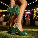 A person outdoors at night wearing sparkling emerald green high-heeled sandals with rhinestone embellishments, holding a matching green rhinestone clutch with a gold chain strap. The background shows blurred string lights and guests in a festive outdoor setting, Obroi