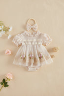 A baby girl's short-sleeve romper in a soft cream/beige color, featuring a textured knit bodice, ruffled collar, lace appliques, and pink floral embroidery on the tulle skirt. It is styled with a matching beige bow headband on a warm beige background with decorative flowers and balls, Obroi