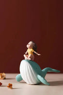 A decorative figurine of a girl in a yellow top sitting cross-legged on a whale, set against a warm brown background, Obroi
