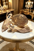 A pair of pointed-toe slingback heels in a champagne pink satin finish, decorated with large crystal-embellished floral embellishments on the toes and heels, shown alongside a matching woven crystal clutch on a white marble table in an ornate gold interior, Obroi