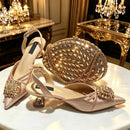 A pair of pointed-toe slingback heels in a champagne pink satin finish, decorated with large crystal-embellished floral embellishments on the toes and heels, shown alongside a matching woven crystal clutch on a white marble table in an ornate gold interior, Obroi