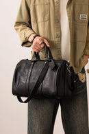 A person wearing a beige button-up shirt and gray jeans holds a black leather duffel bag with top handles and a shoulder strap against a plain light background, Obroi