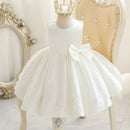 A white sleeveless flower girl dress with a large bow on the skirt, displayed on a mannequin in an ornate room with a gold crown accessory, Obroi